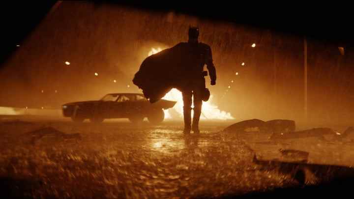 Batman arka planda bir ateşle yağmurlu bir sokakta kameraya yaklaşıyor.