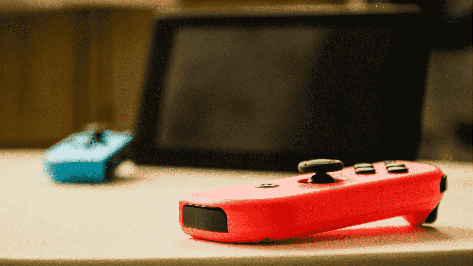 Ön planda bir masada neon kırmızısı bir Joy-Con, Nintendo Switch ekranı ve arka planda masanın üzerinde duran mavi Joy-Con, biraz odak dışı