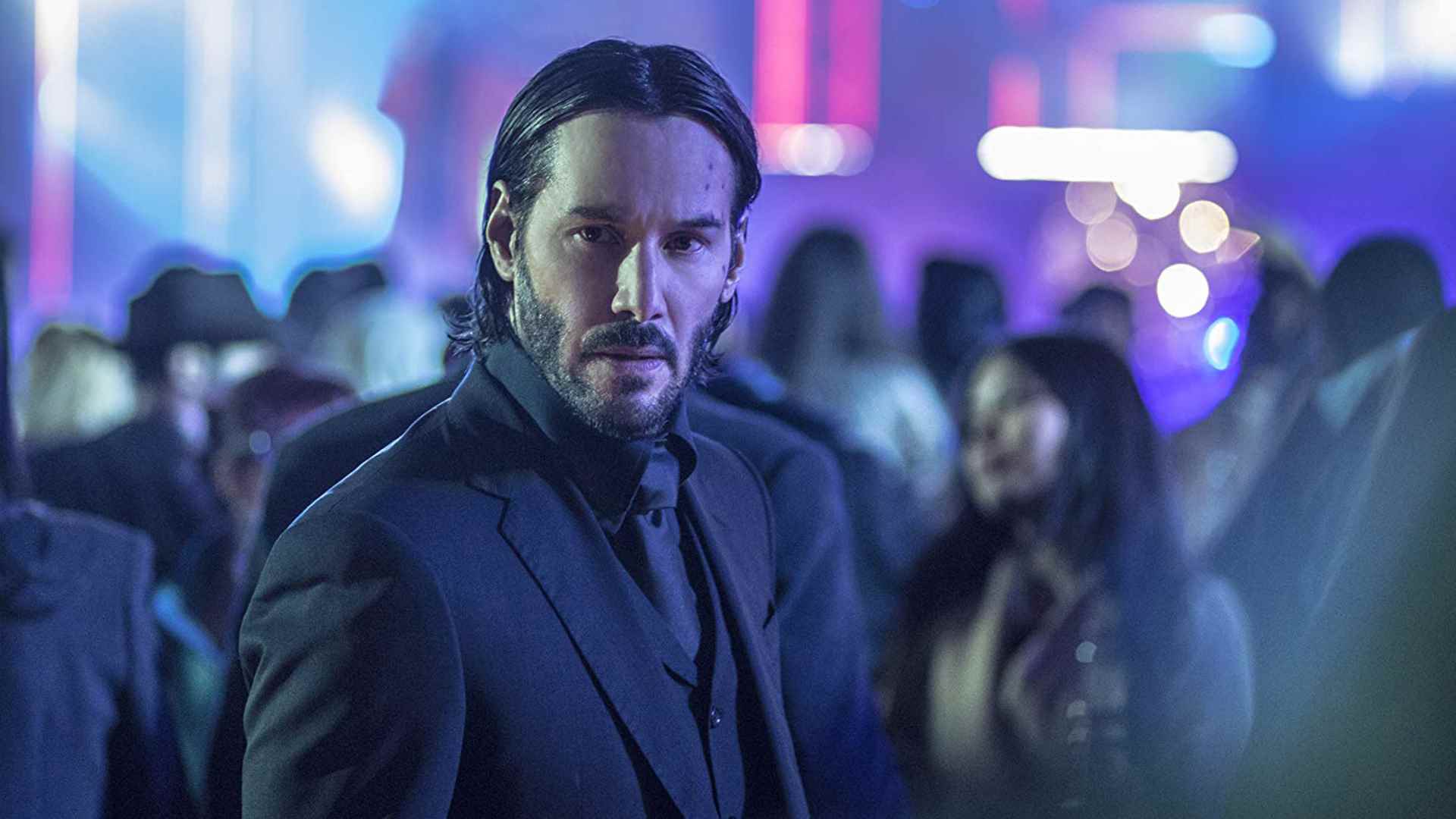 Keskin bir takım elbiseli John Wick, filmlerinden birinde neon bir ortamda birini arar.