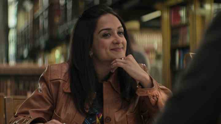 You 4. sezondan Nadia bir kütüphanede masada oturuyor, eli çenesinde, gülümsüyor.