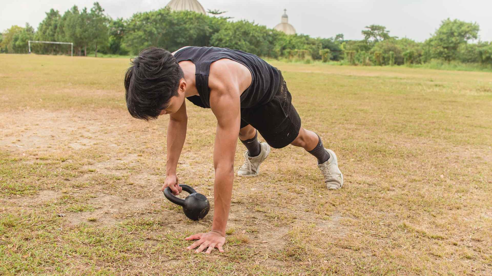 Tahta pozisyonundaki çimenlerin üzerinde bir kettlebell'i bir taraftan diğerine hareket ettiren adam