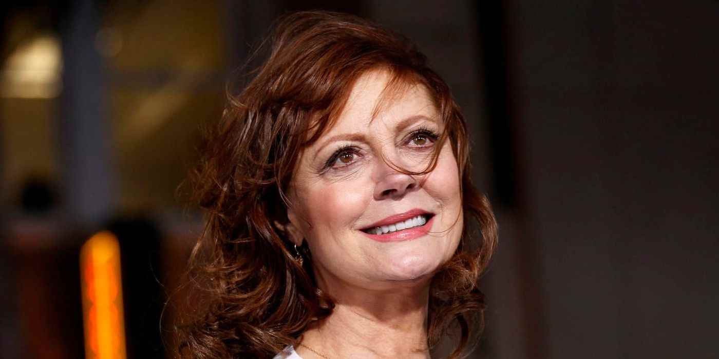 Susan Sarandon, yüzündeki saçlarla yukarı doğru bakarak gülümsüyor.