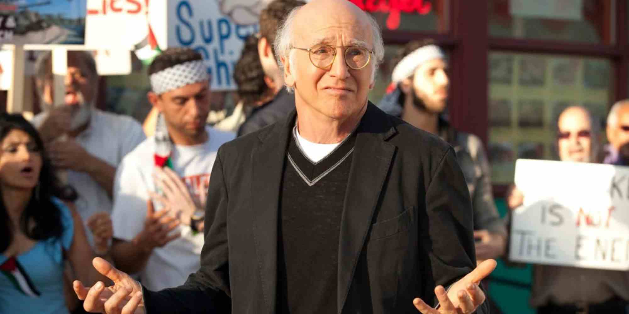 Larry David, Curb Your Enthusiasm'daki bir protestonun önünde omuz silkiyor.