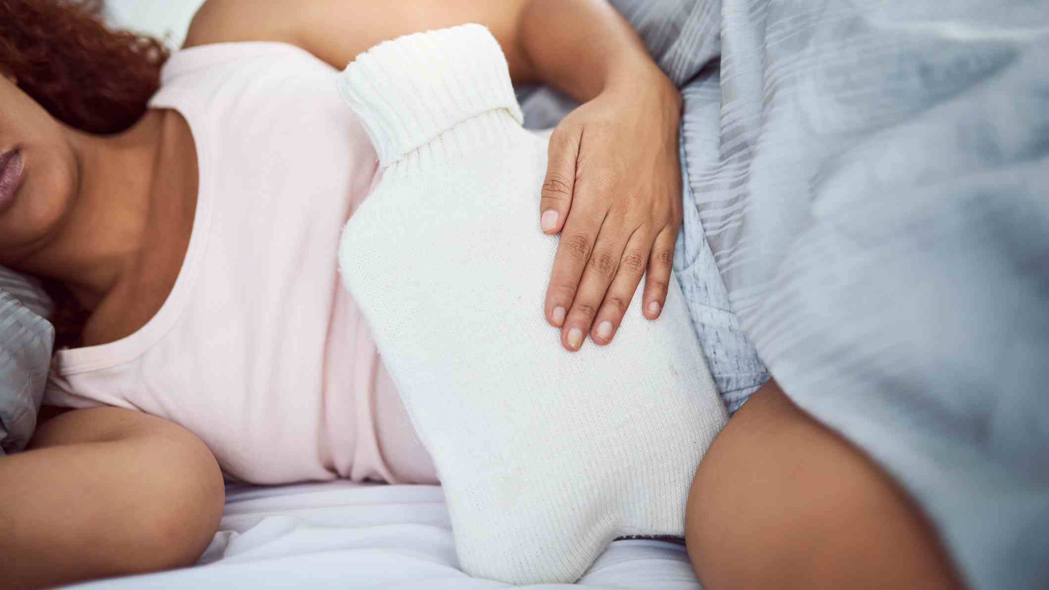 Pembe uyku yeleği giyen bir kadın elinde beyaz sıcak su şişesiyle yatakta yatıyor.