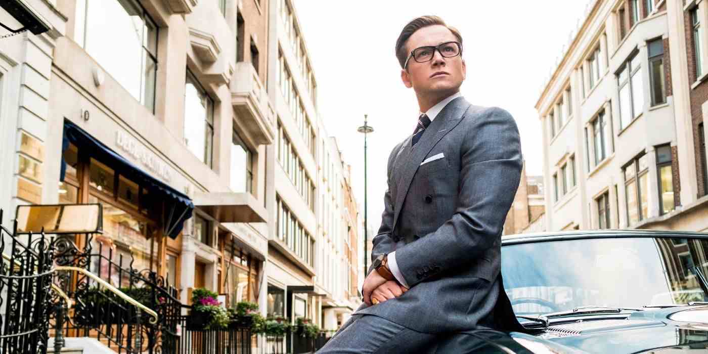 Kingsman: The Golden Circle'da iyi giyimli ve ağırbaşlı bir Eggsy, vintage yeşil bir otomobilin tepesinde oturuyor ve yan tarafa bakıyor.