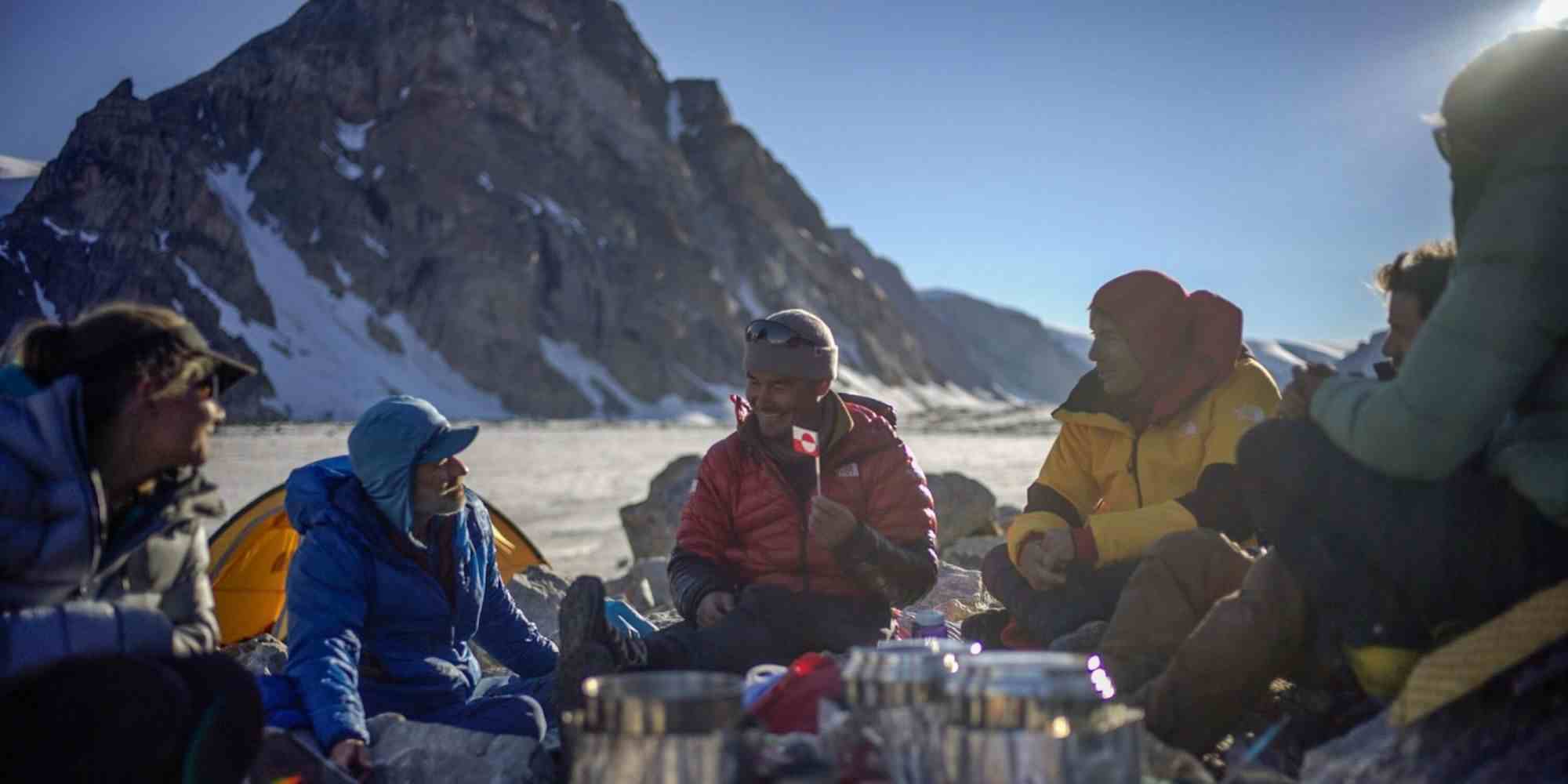 Ağır paltolu ve şapkalı bir grup kaşif ve dağcı, Alex Honnold'la birlikte Arctic Acsent'teki bir kaya duvarının önünde kardaki kamplarında oturuyor.