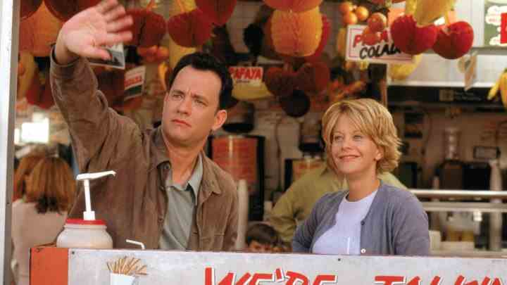 Tom Hanks, Meg Ryan'ın yanında dururken elini kaldırıyor.