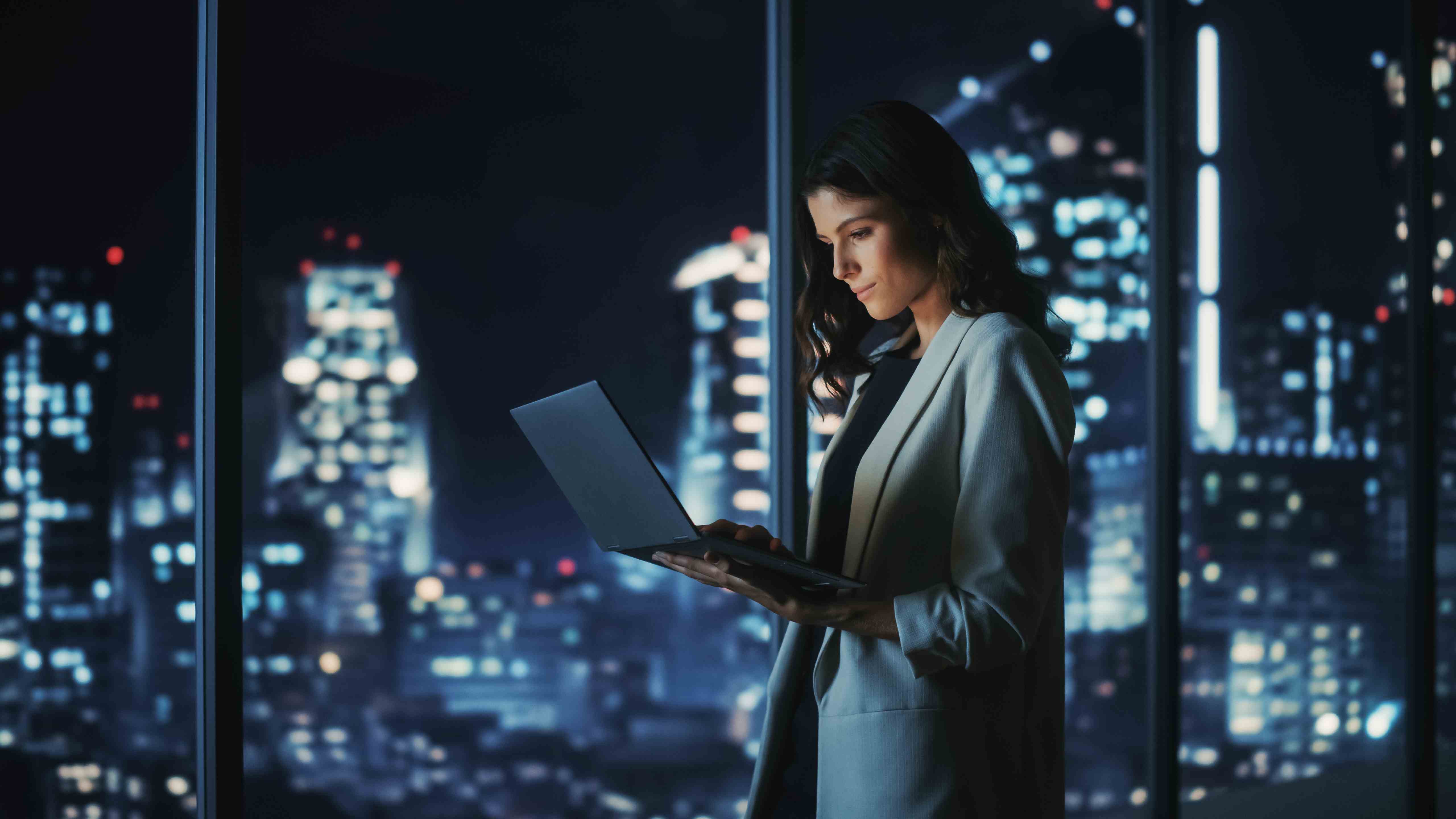 Gece vakti, arka planında şehir manzarası olan bir odada duran kadın, elinde bir dizüstü bilgisayar tutuyor ve tek eliyle kullanıyor.